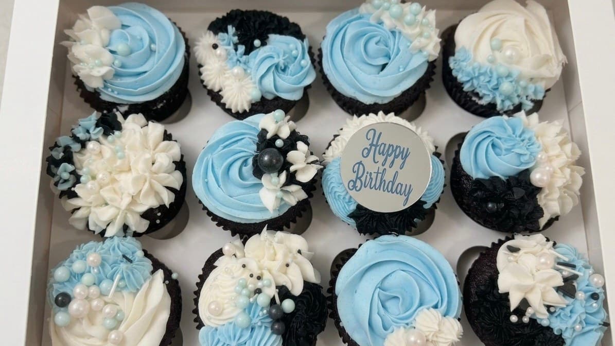 A boxed dozen of chocolate cupcakes iced in blue and white, with sugar pearls and a Happy Birthday topper